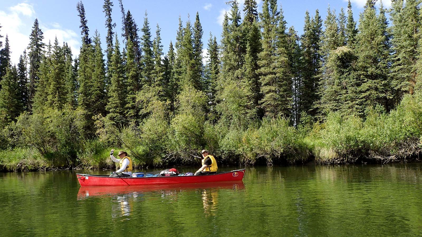 Two people in a red canoe on a calm green river with dense pine forest behind them