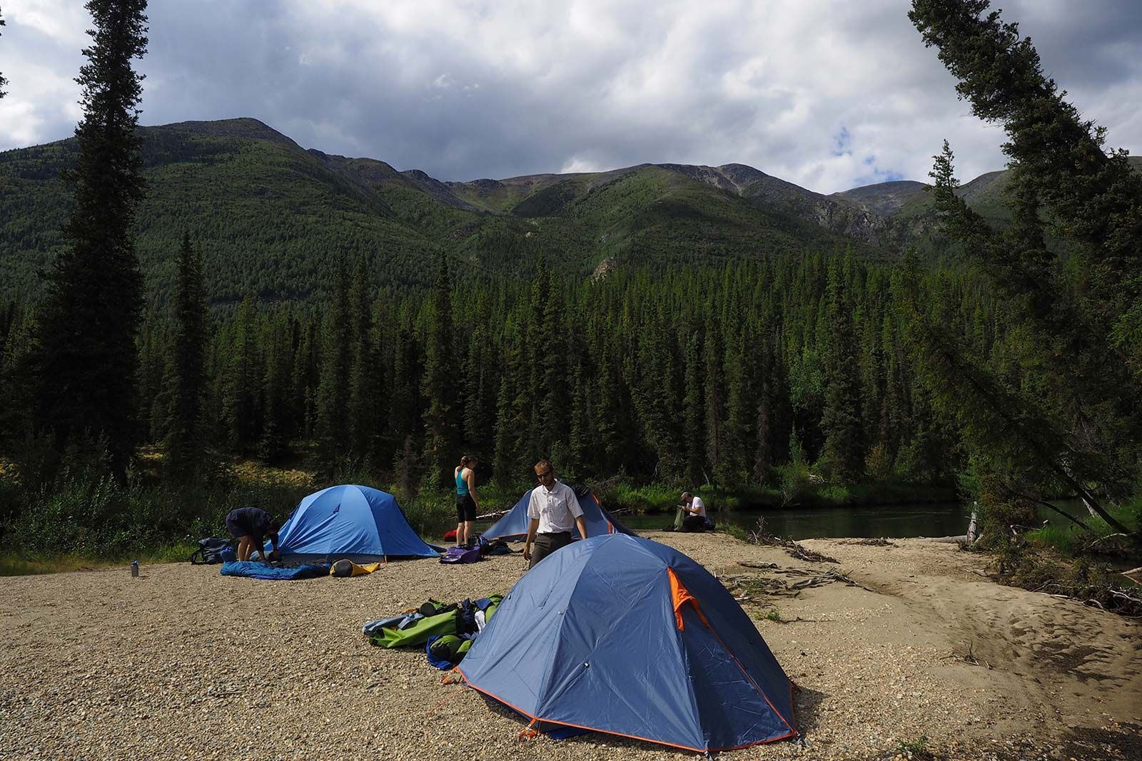 Campers near blue tents on a rocky mountain campsite by a forested lake and peaks.