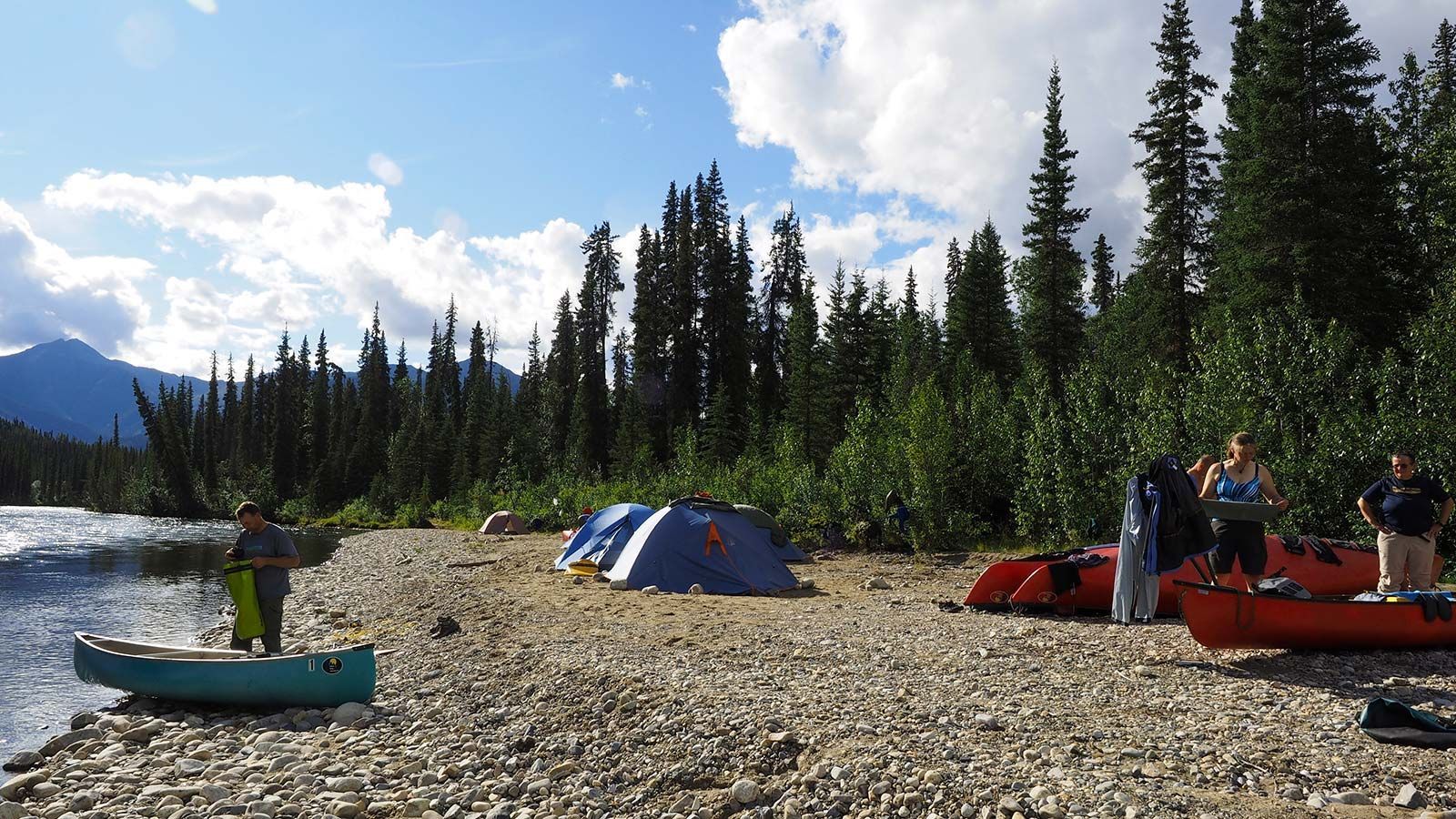 Campers on a rocky riverbank with tents, red canoes, and a forested mountain backdrop