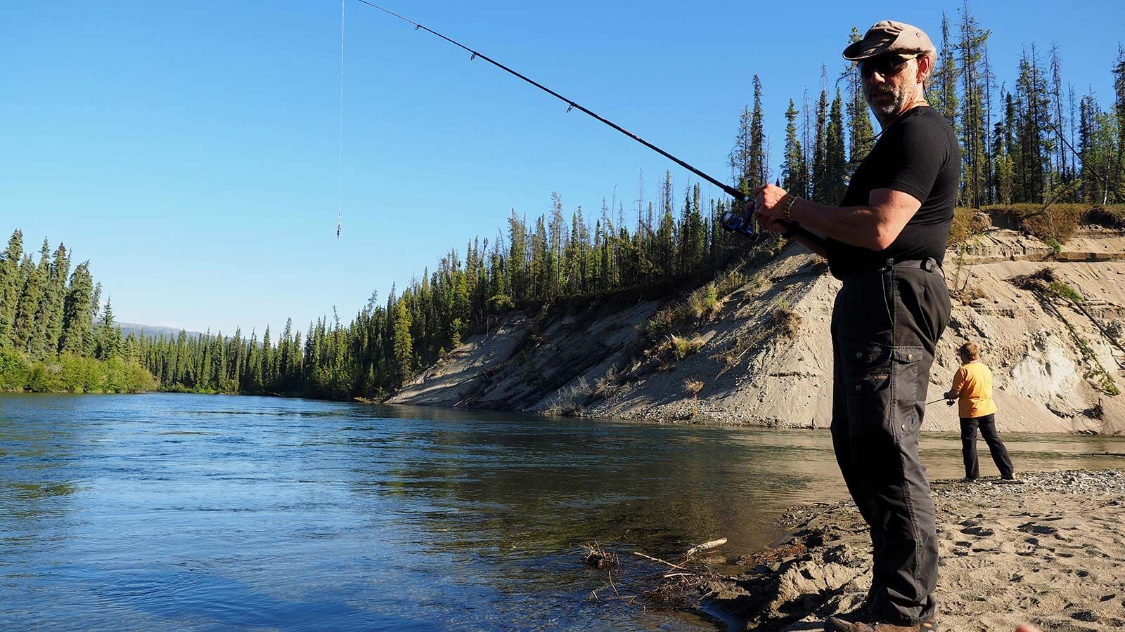 Angler in black fishing by a riverbank, with another person in yellow farther back under a clear blue sky.