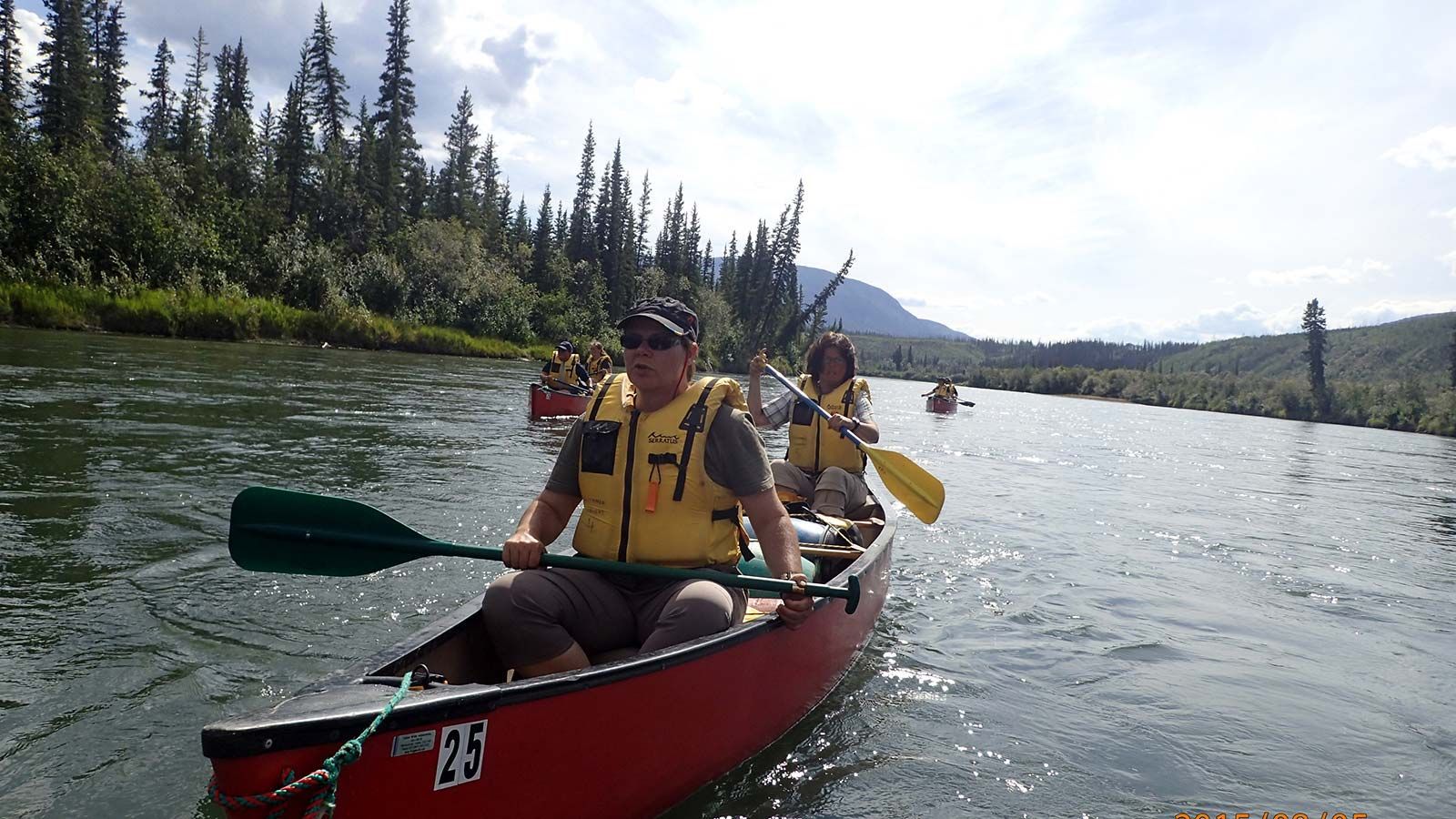 Two people canoeing on a river, with evergreen trees and mountains in the background