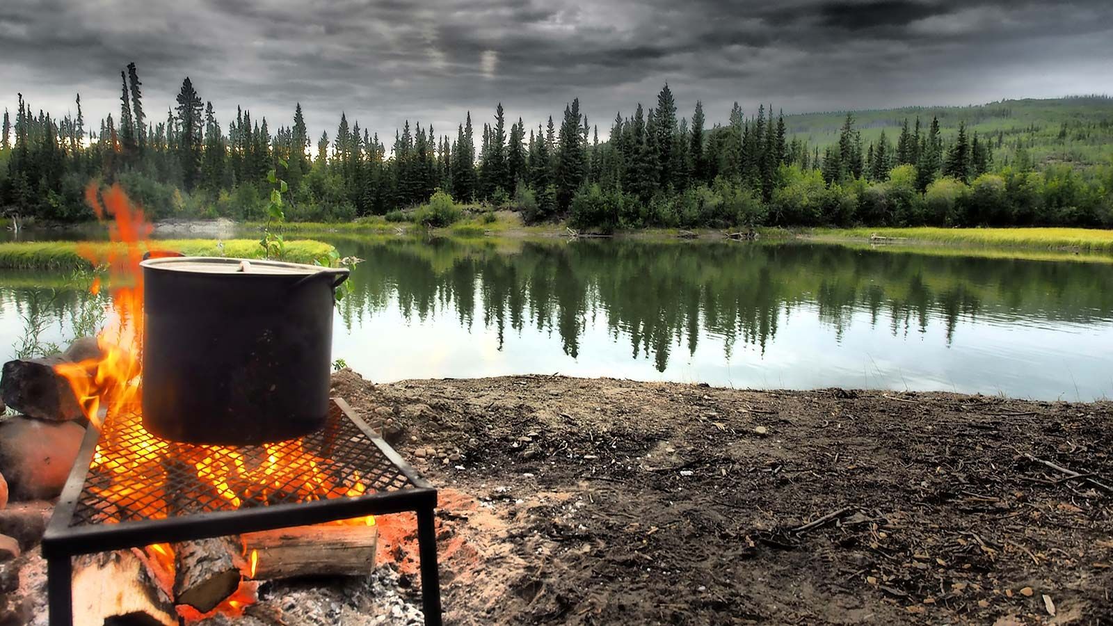 Campfire cooking pot beside a calm lake with pine forest under a cloudy sky