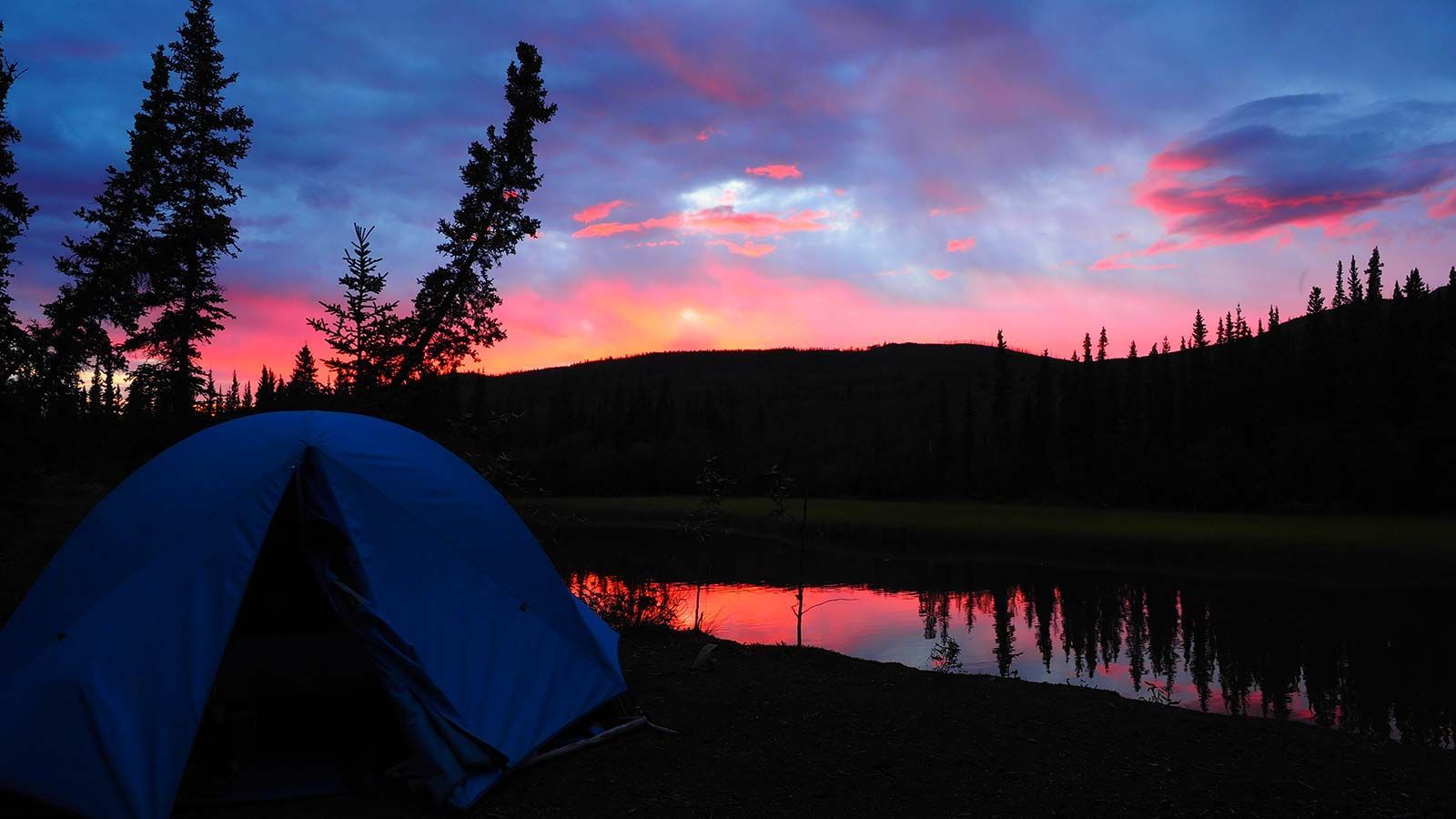 Blue tent by a lake at sunset, with pink clouds and dark trees silhouetted against the sky