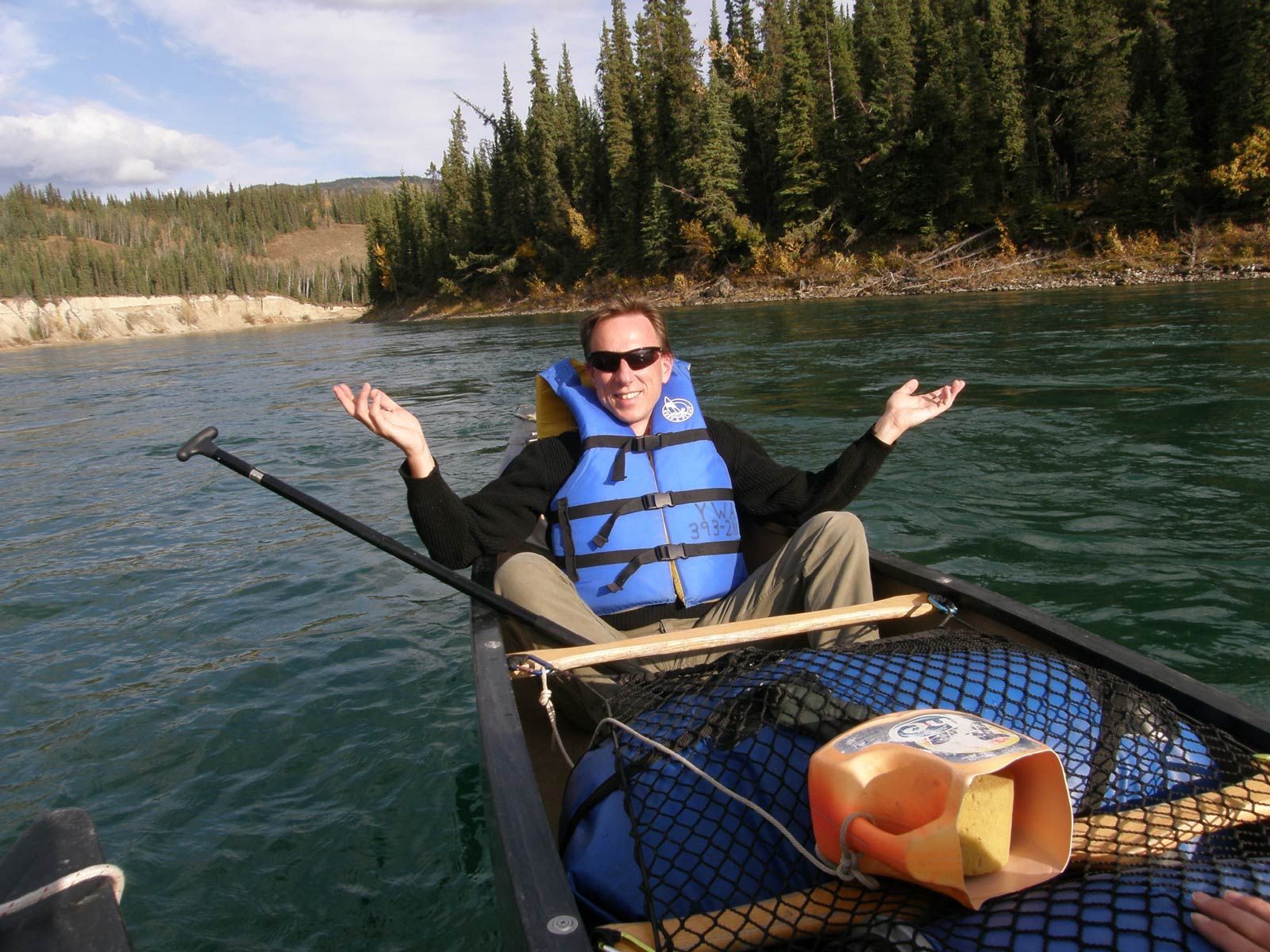 Person in a blue life vest kayaking on a river with raised arms, forested banks in the background