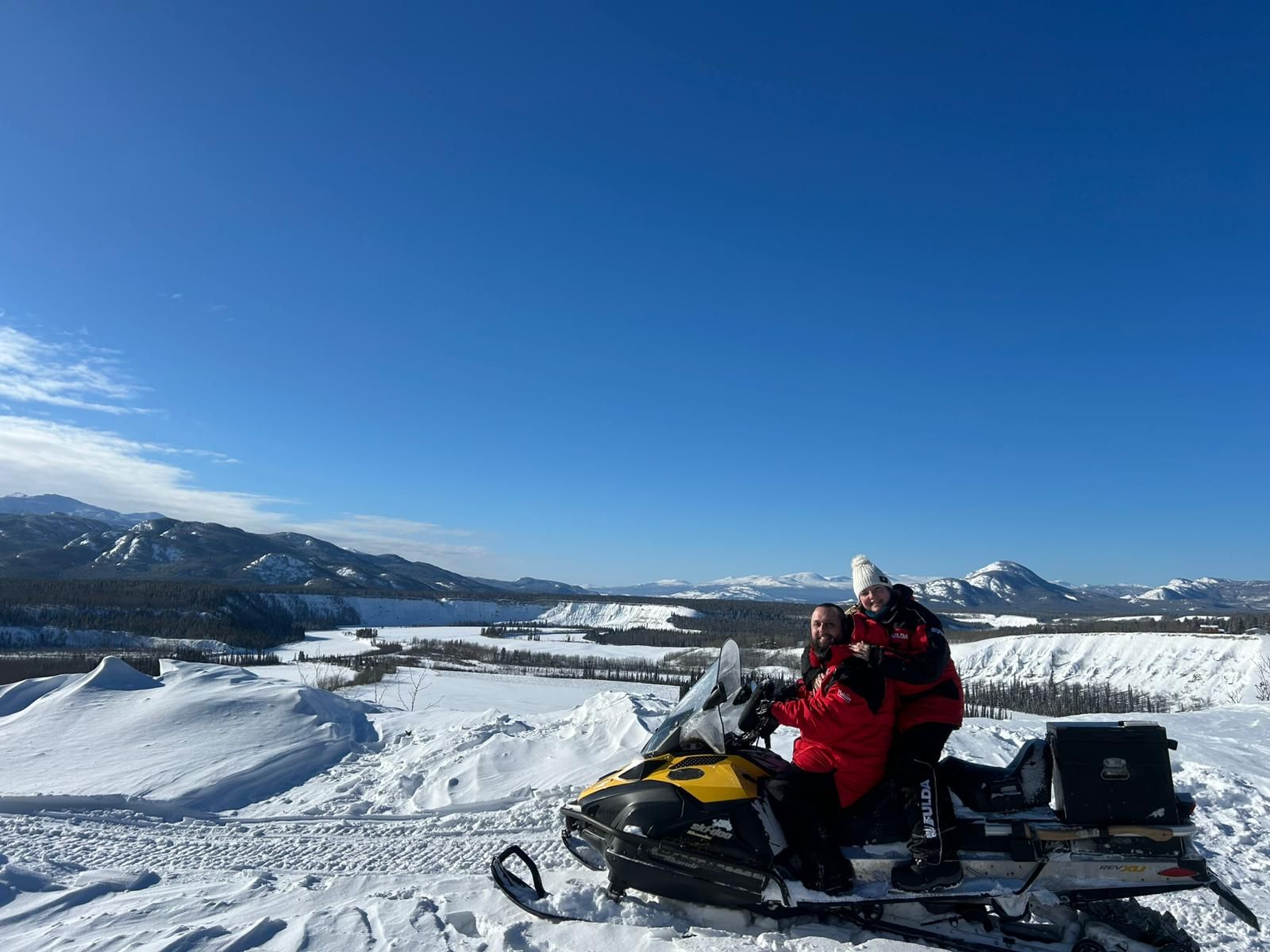 Snowmobilers posing on snowy terrain with black and yellow snowmobiles under a blue sky