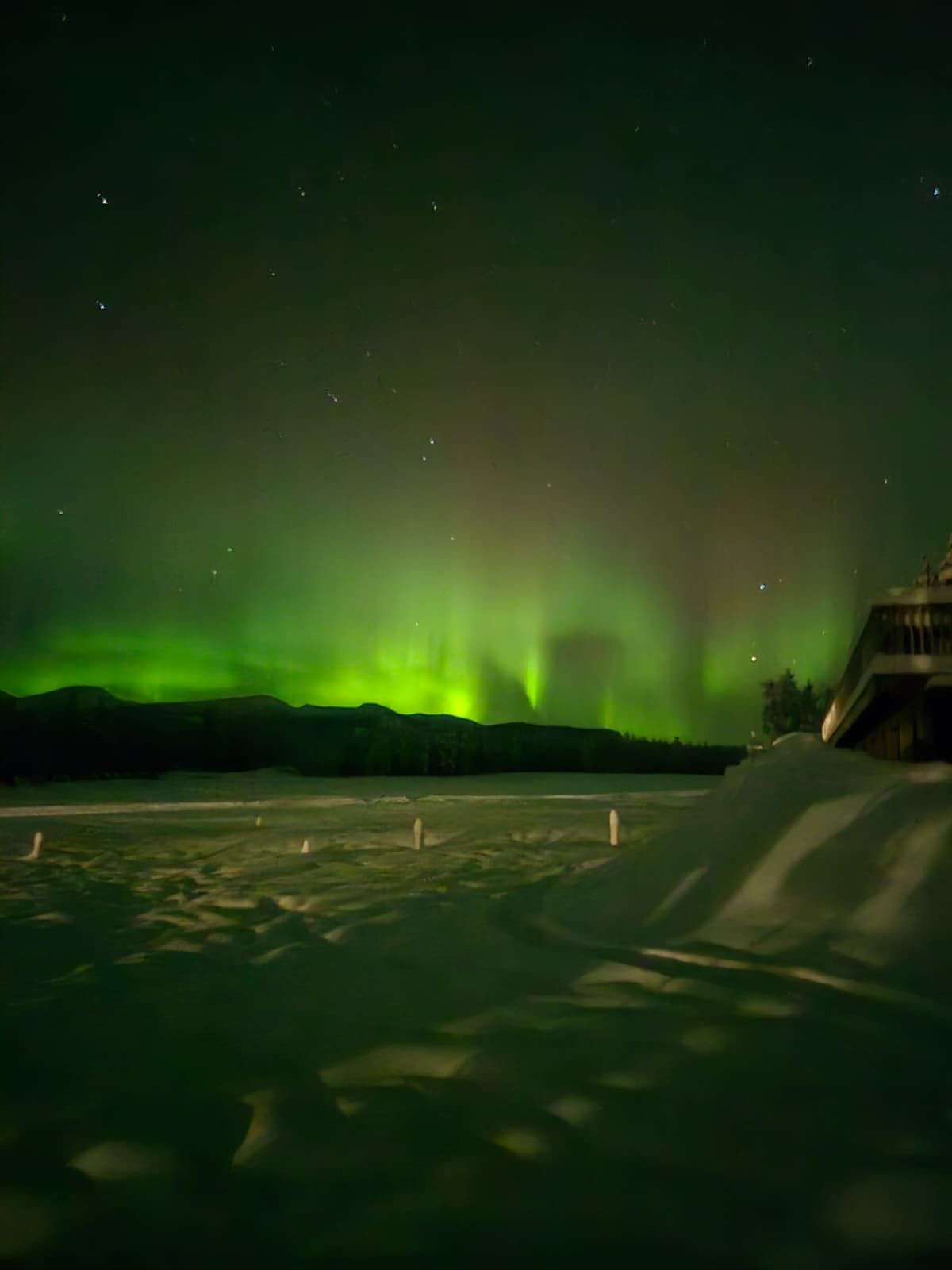 Green aurora over snowy landscape at night with dark mountains and a lit building on the right