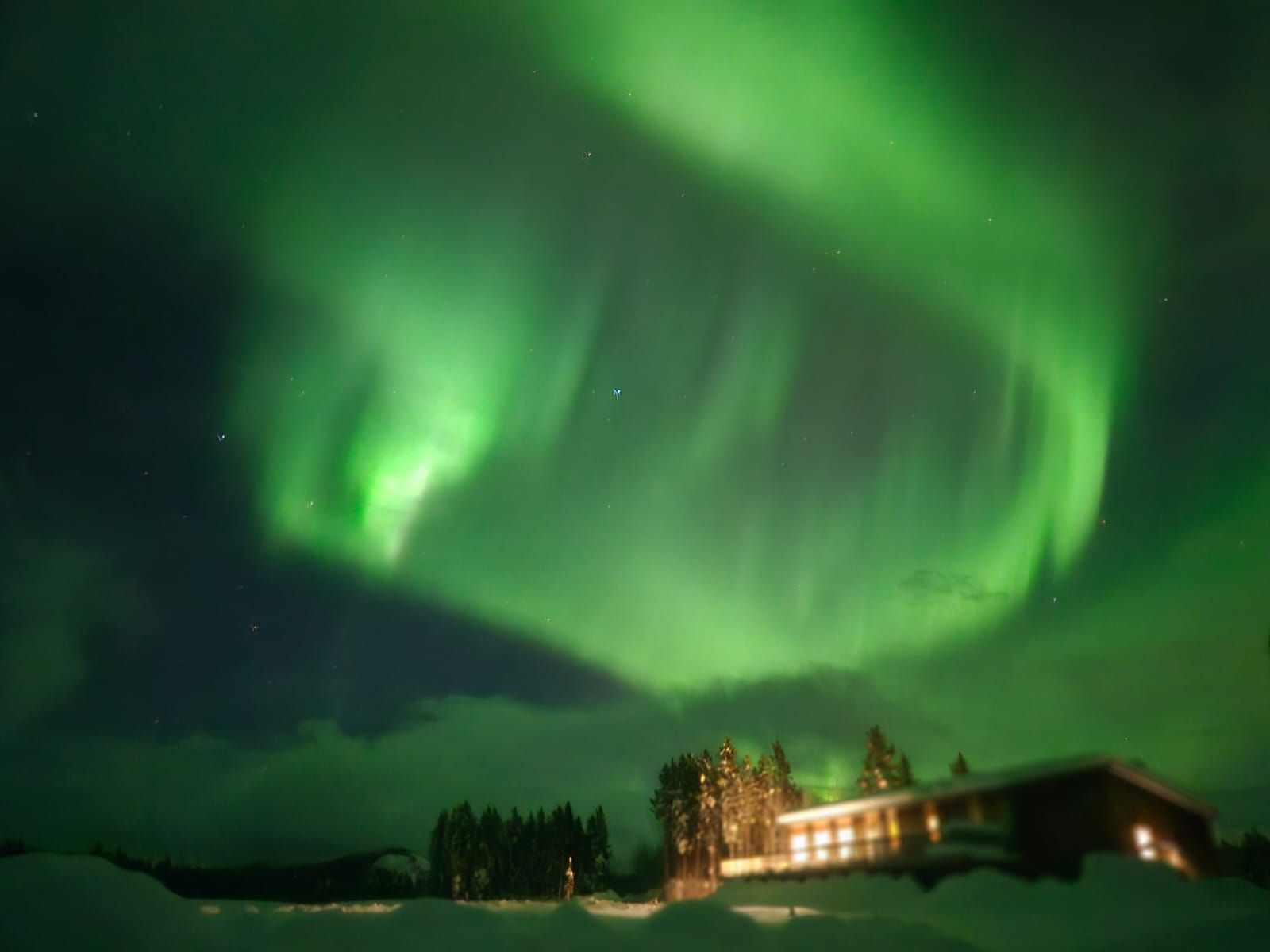 Green aurora borealis over a snowy landscape with a lit lodge at night