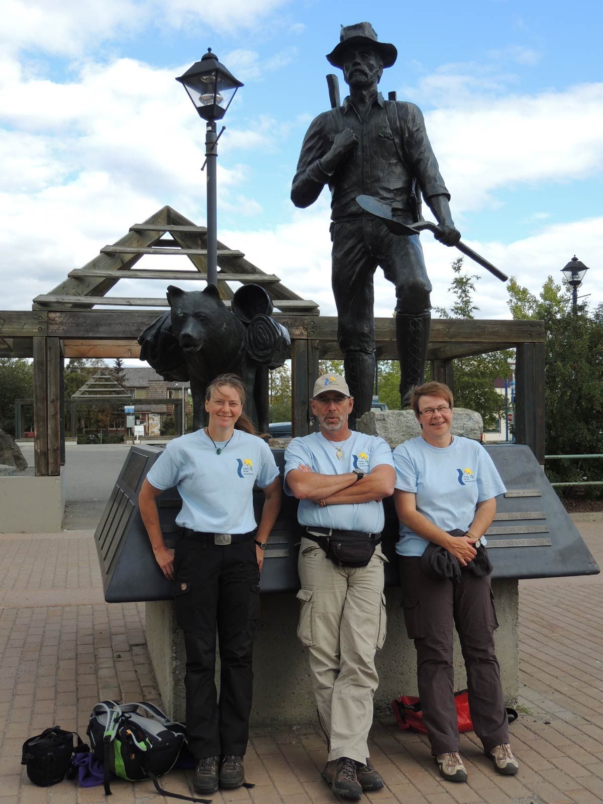 Three people posing beside a bronze statue in a park, with a lamp post and wooden structure behind them.