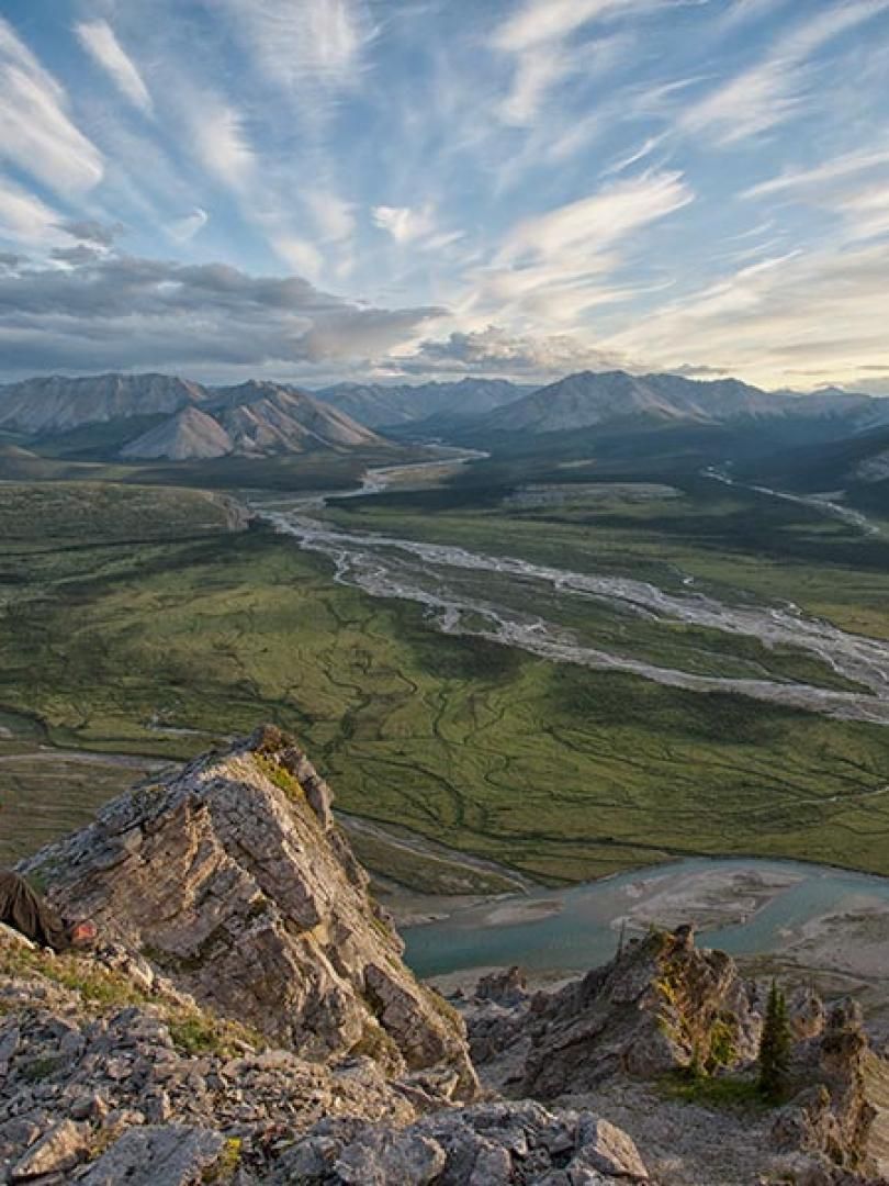 Rocky mountain overlook with a winding river, green valley, and dramatic cloudy sky at sunset