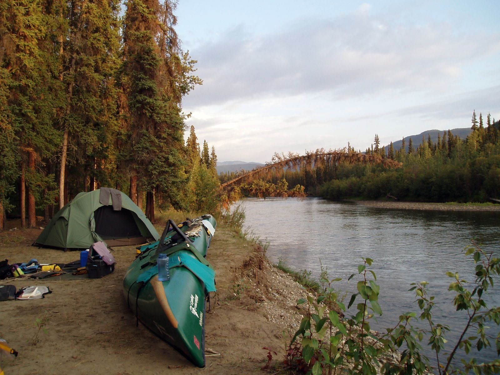 Green tent and canoe on a riverbank beside a calm river at dusk, with forested hills in the background