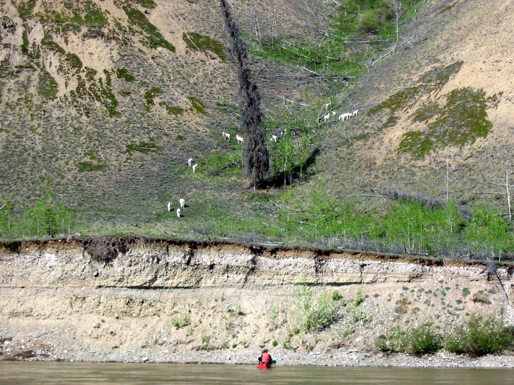 Steep rocky hillside above a riverbank, with a narrow stream and two small hikers near the water.