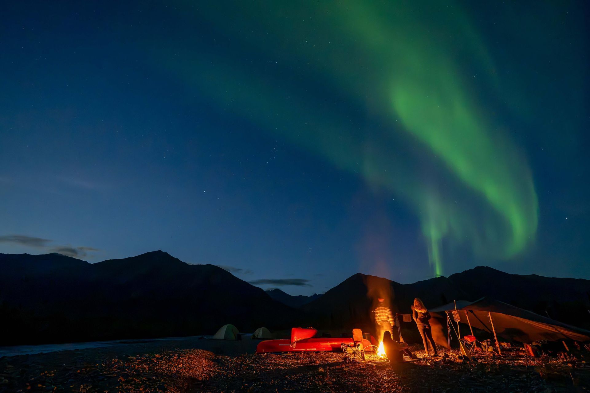 Campfire on a rocky shore under green northern lights, with dark mountains silhouetted at dusk.