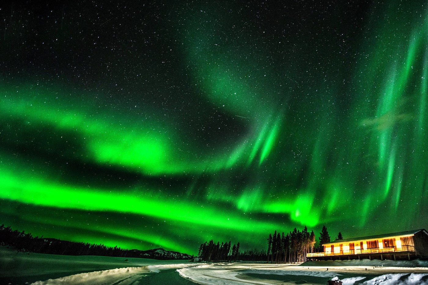 Green aurora borealis over a snowy landscape with a lit cabin at night