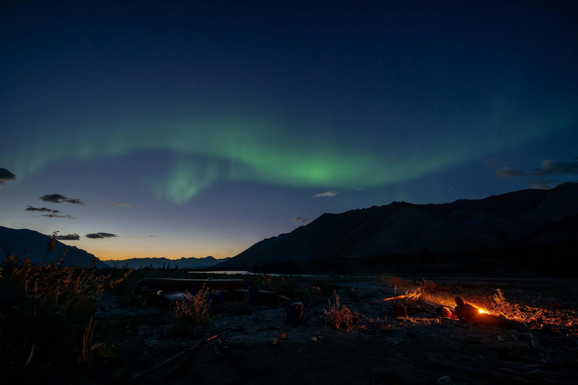 Northern lights over a dark mountain valley at dusk, with a small campfire glowing in the foreground