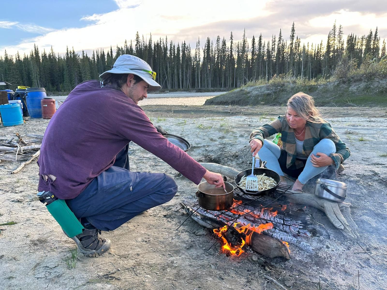 Two campers cooking over a campfire by a lake, with trees and gear in the background.