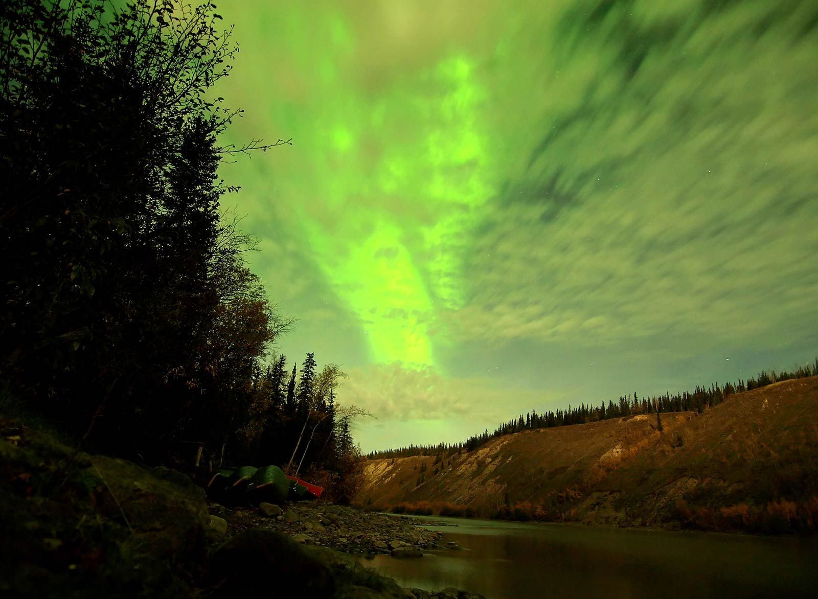 Green aurora glowing over a dark forest and river at dusk