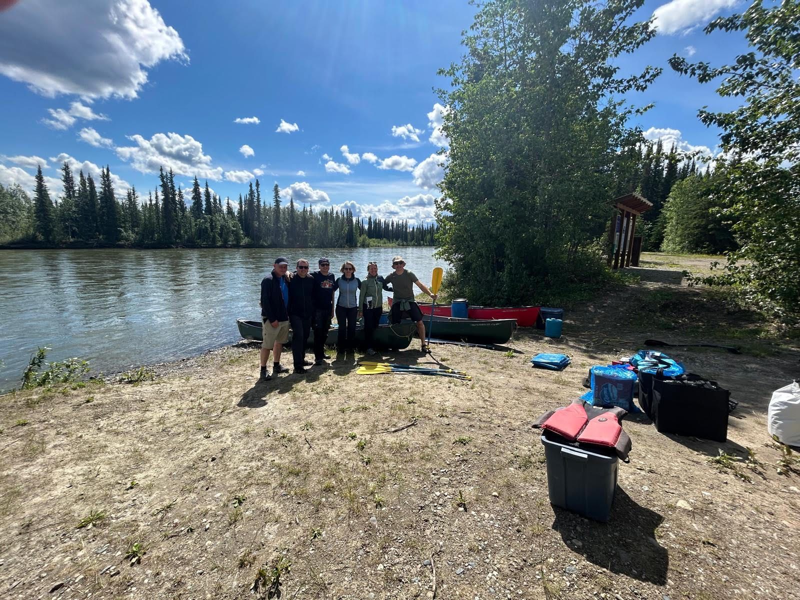 People posing by a lakeshore with trees, kayaks, and gear under a bright blue sky.