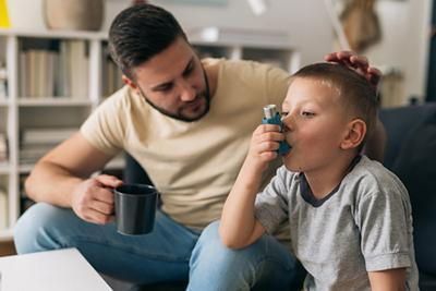 A man and a boy are sitting on a couch and the boy is using an inhaler.