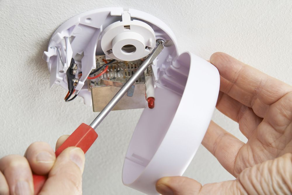 Person using a screwdriver to open a white smoke detector on a ceiling. — Paul Wiedman Electrical in Harristown, QLD