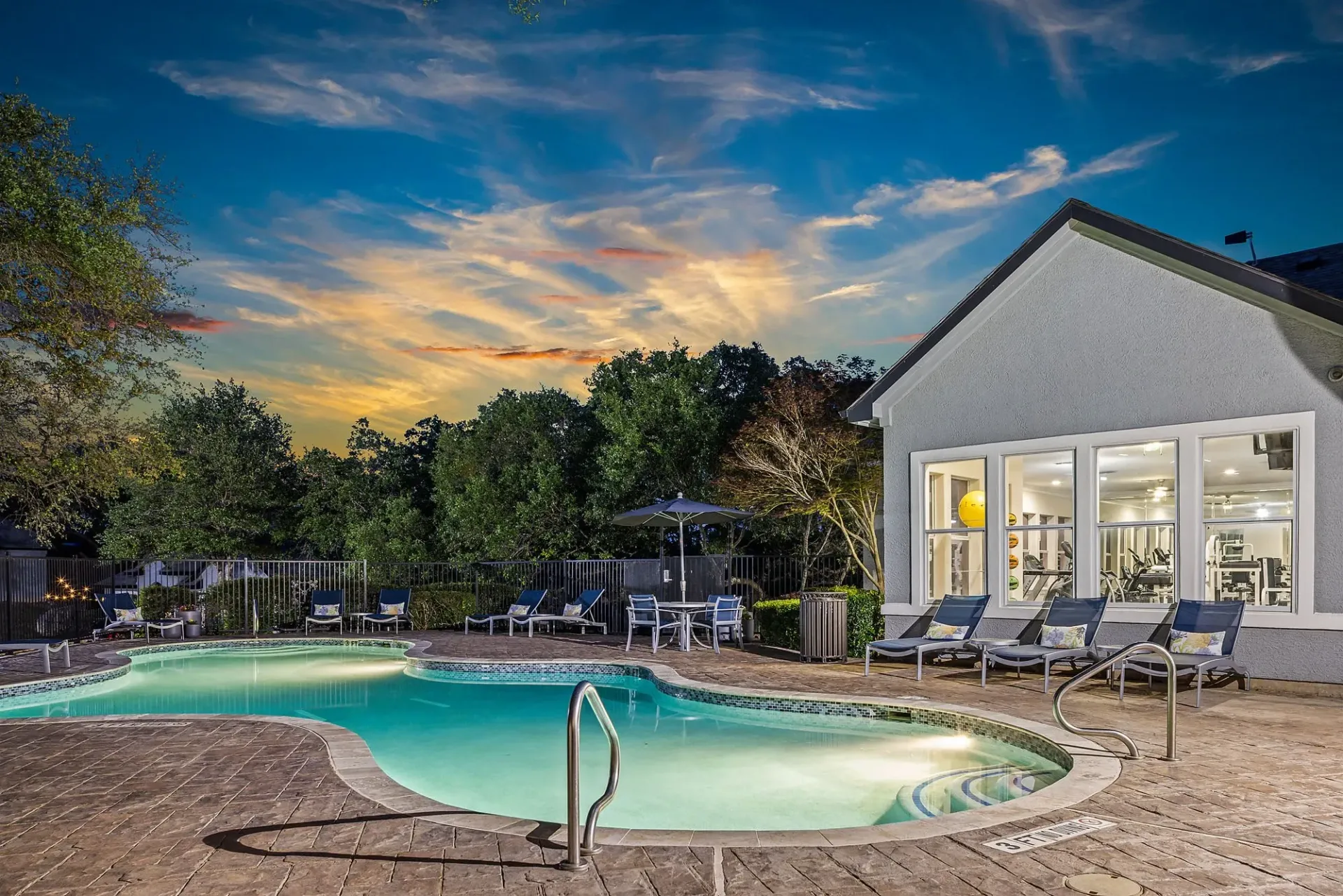 A brightly lit swimming pool and adjacent fitness center visible through large windows, set against a twilight sky.