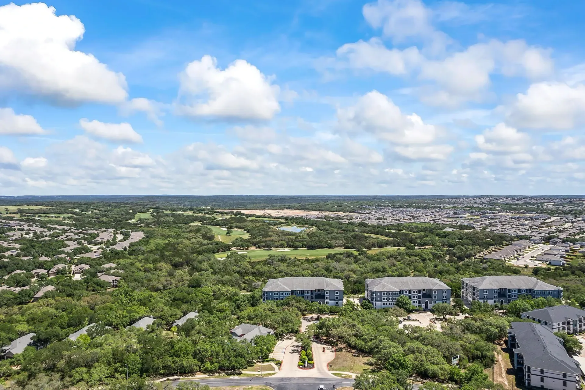 Aerial view of apartment buildings surrounded by lush greenery and a distant golf course under a cloudy blue sky.