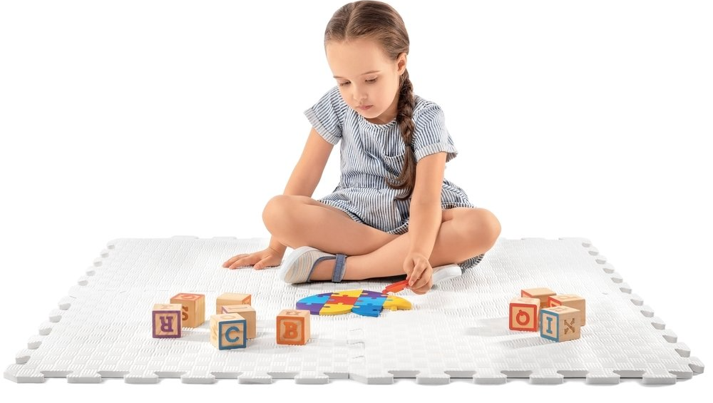 A young girl with a braid sits on a white interlocking foam mat playing with alphabet blocks and colorful shapes.