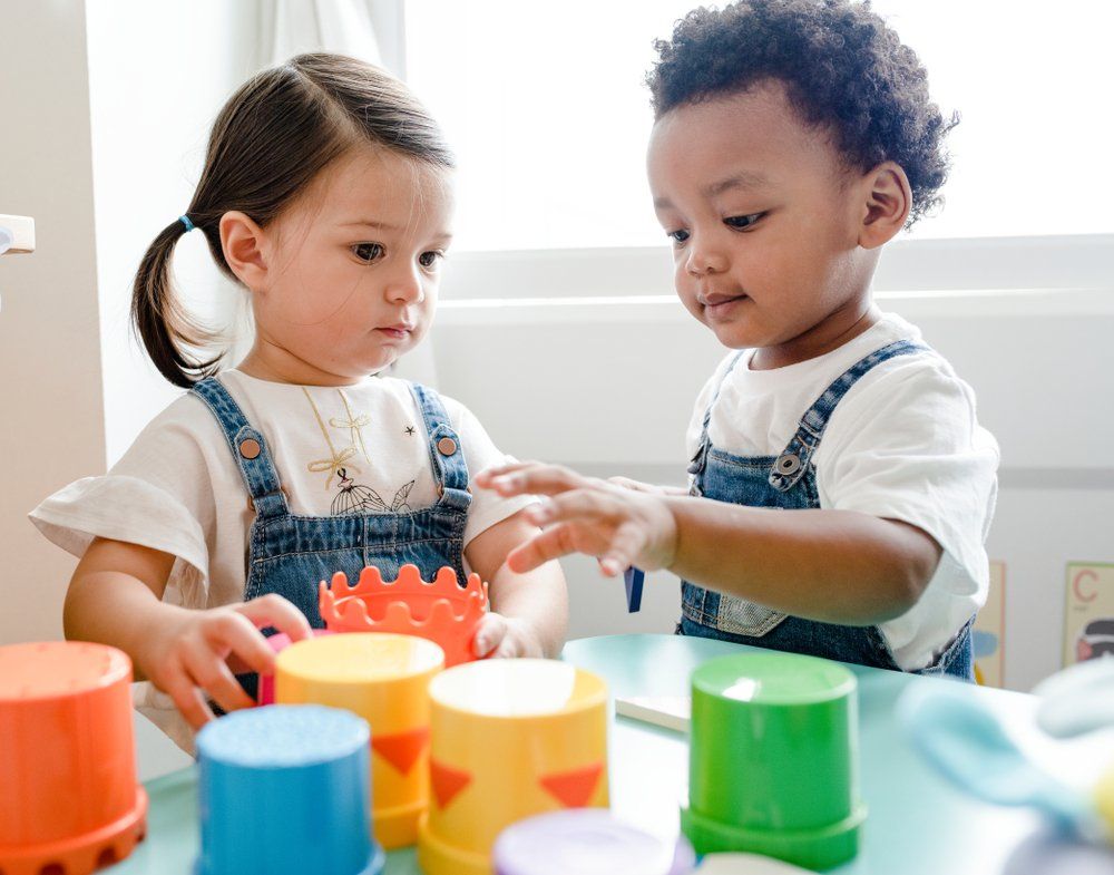 Two toddlers playing with colorful stacking cups at a table in a bright room