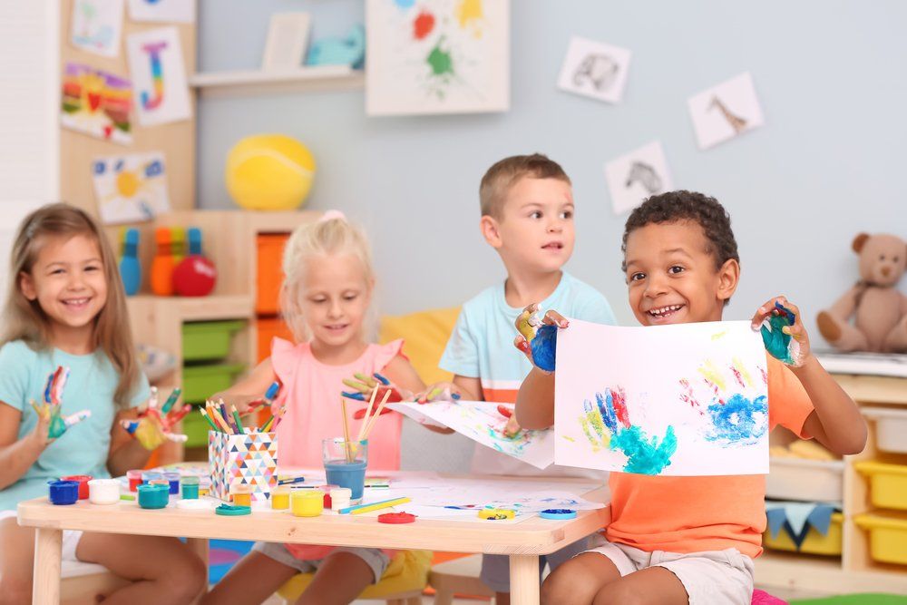 Children painting at a classroom table with colorful art supplies and cheerful drawings.