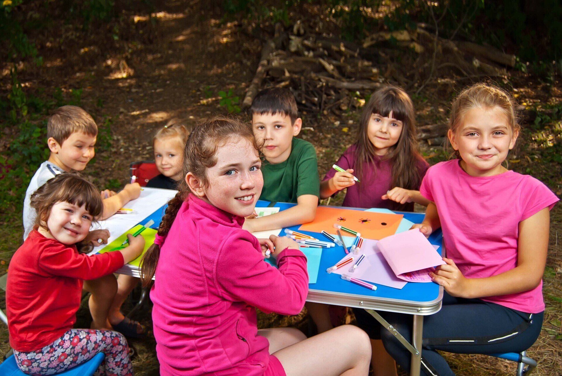 Children gathered outdoors at a small table, drawing and crafting with colorful paper and markers.