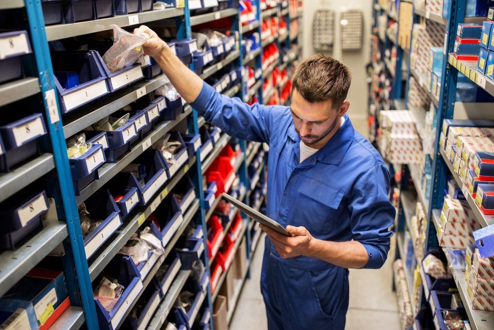 Man in blue jumpsuit, holding tablet, searches a shelf of parts in a warehouse.