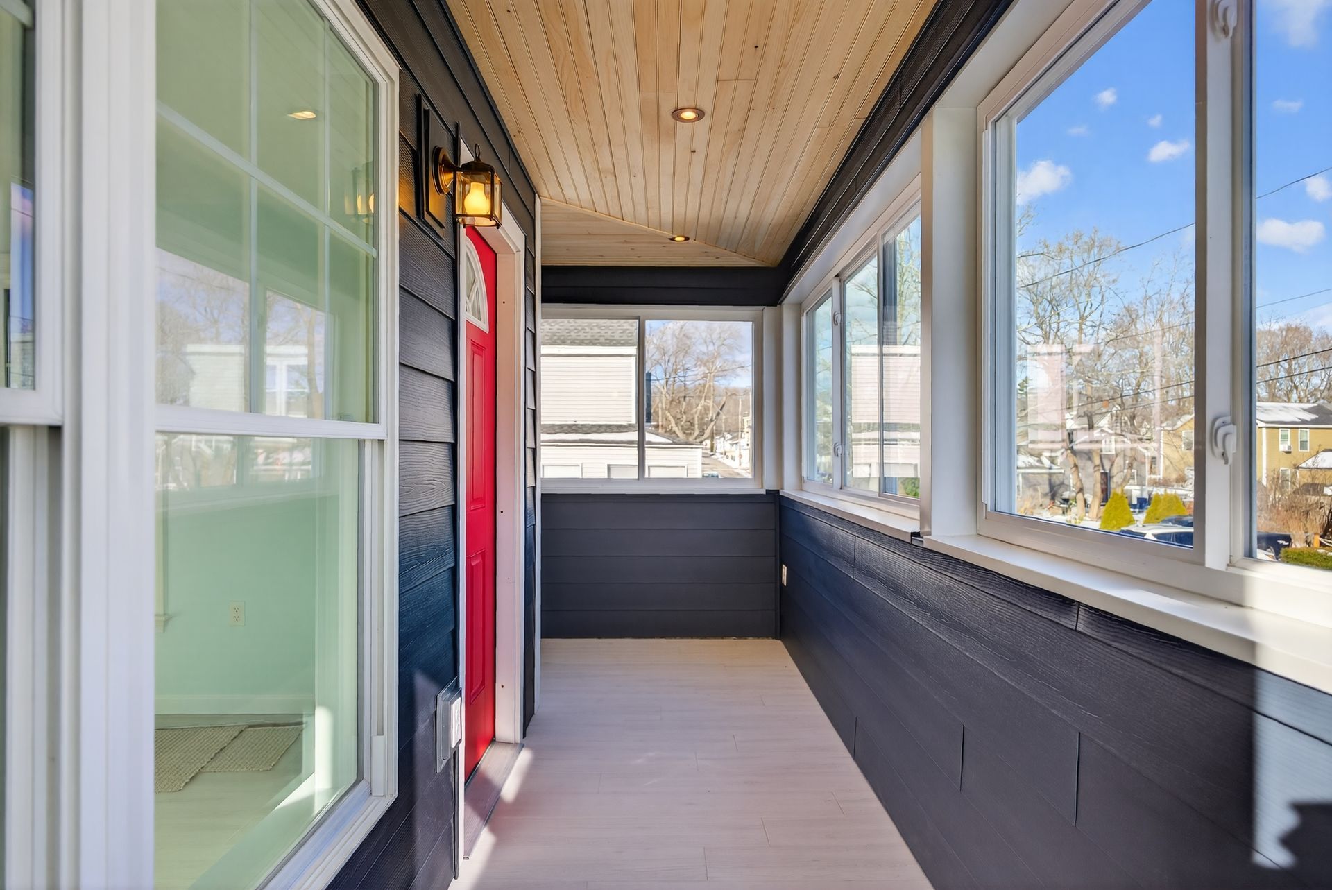 Covered porch with dark siding, white trim windows, red door, wood ceiling, and light-colored floor.