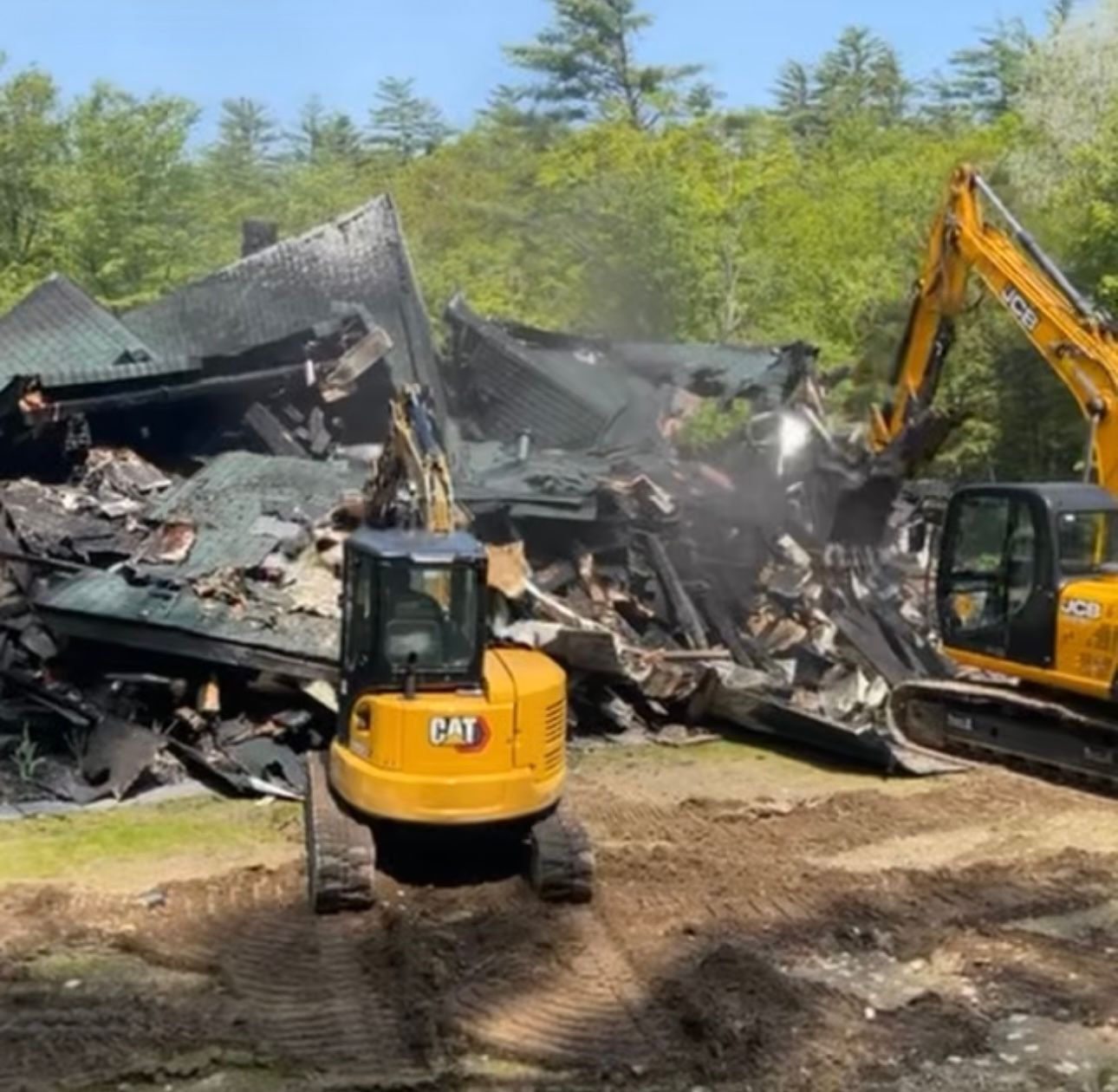 Yellow excavators clear a collapsed, charred building under a blue sky.