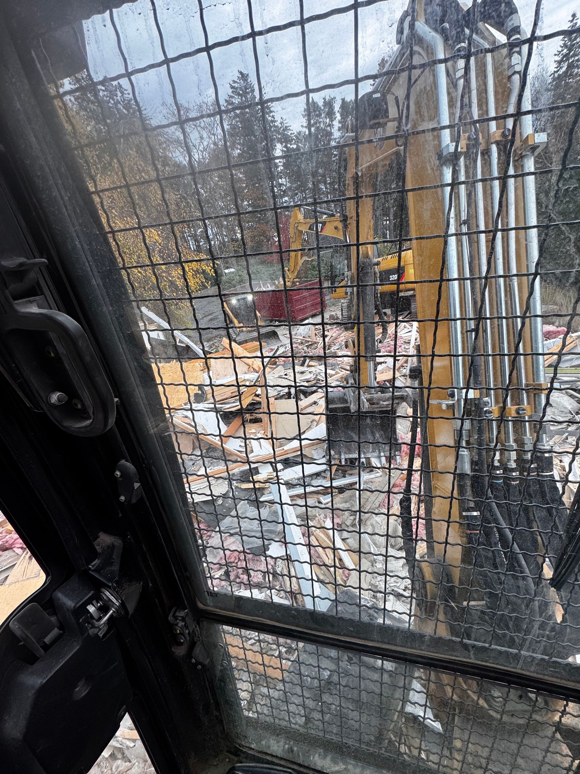 Construction site seen through excavator cab safety grid, with rubble and yellow machinery nearby