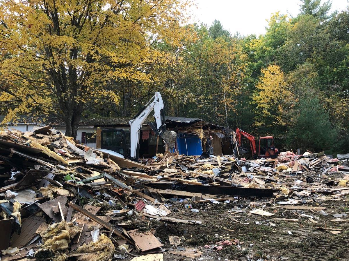 Demolition site with rubble piles, an excavator, and autumn trees in the background