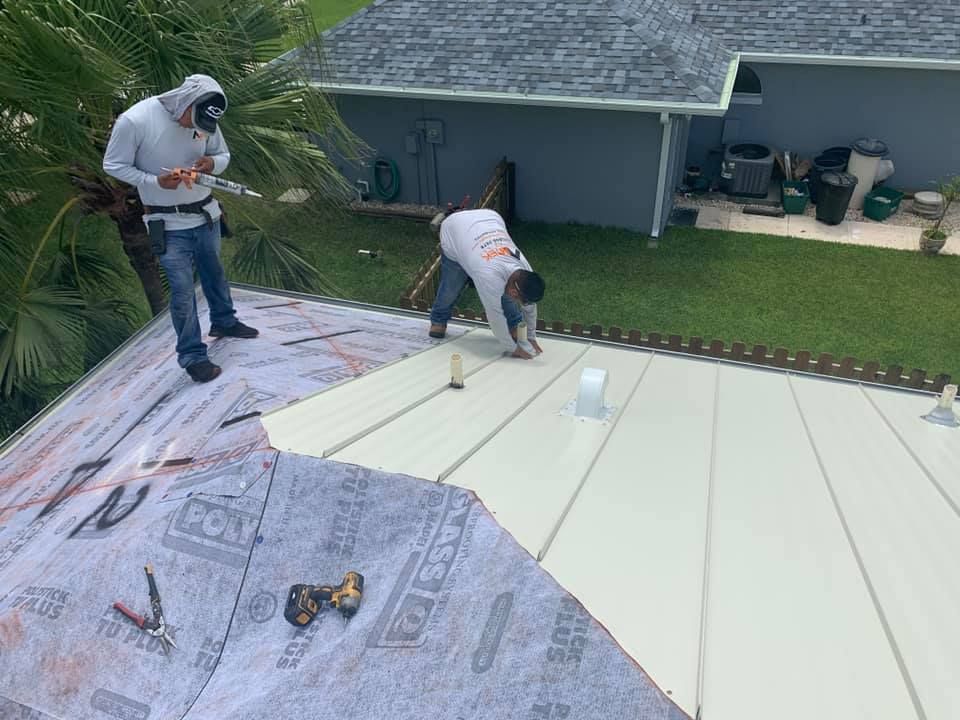 Two workers installing a white roof panel on a house roof, with house and yard in the background