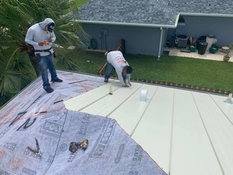 Two workers install roofing panels on a house, with underlayment exposed on one side.