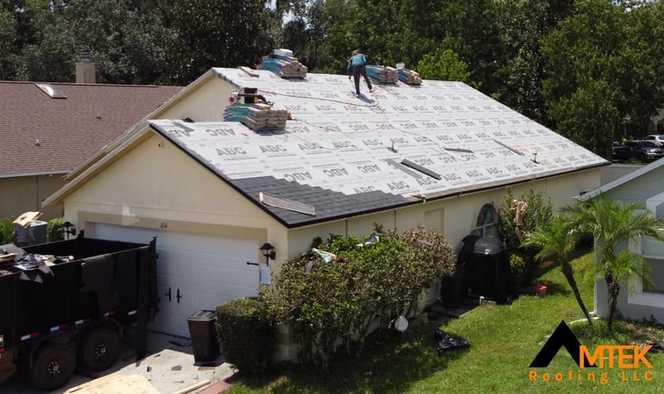 Roofers installing new shingles on a light-colored house roof in a suburban yard.