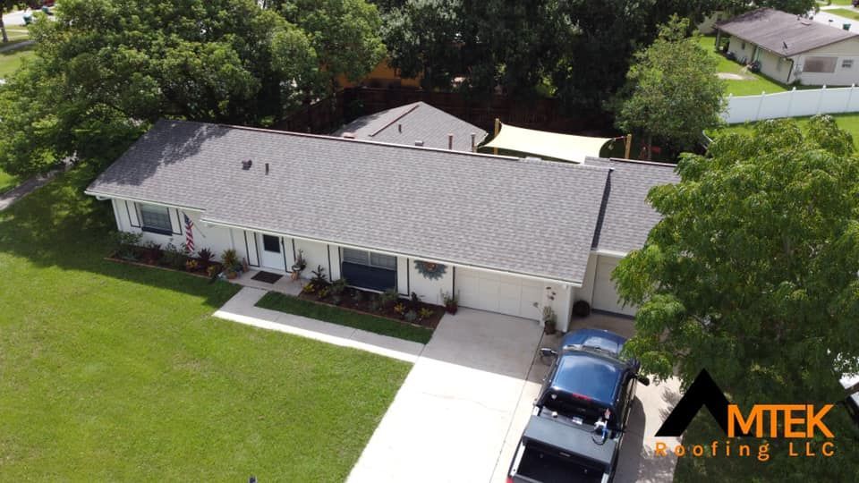 Aerial view of a single-story house with a gray roof, driveway, and green lawn surrounded by trees.