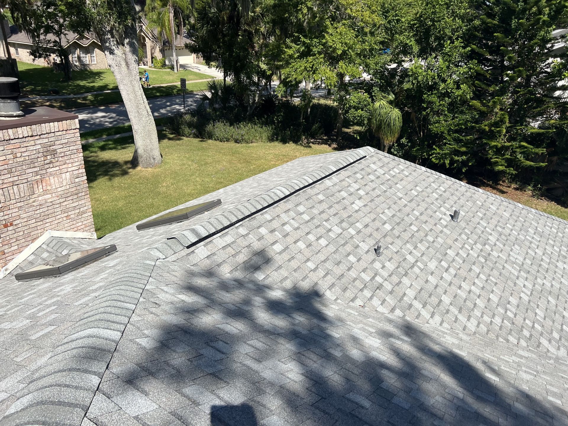 Gray shingle roof with sunlight and tree shadows beside a brick chimney, viewed from above
