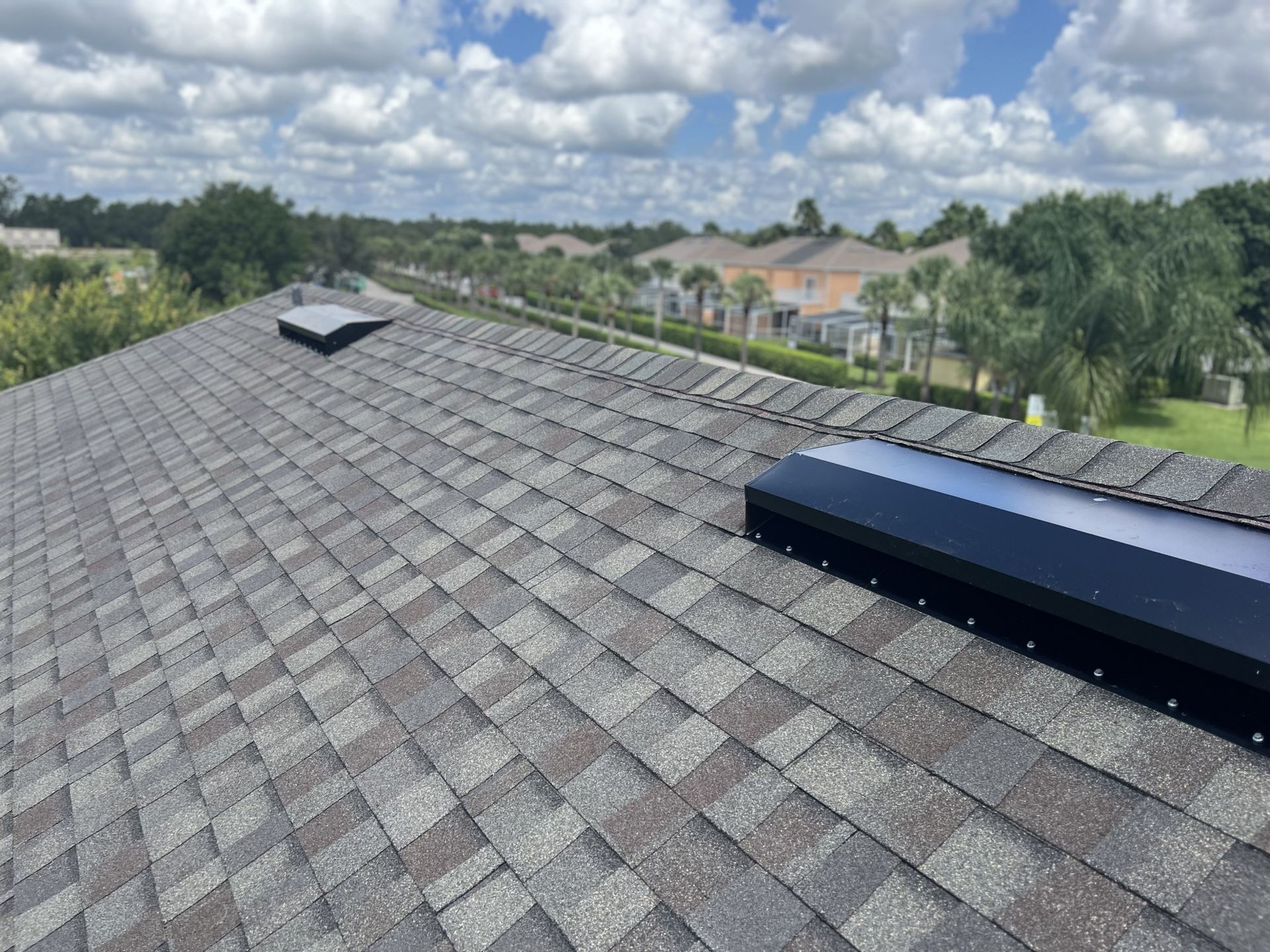Solar panel arrays on a shingled rooftop under a cloudy sky, with trees and houses in the background