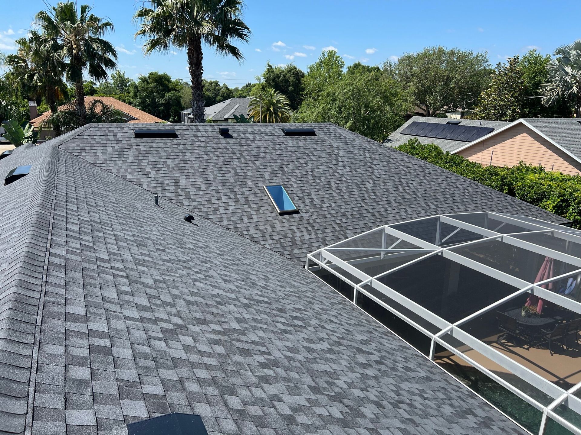 Black shingle roof with skylights and solar panels under clear sky, surrounded by palm trees