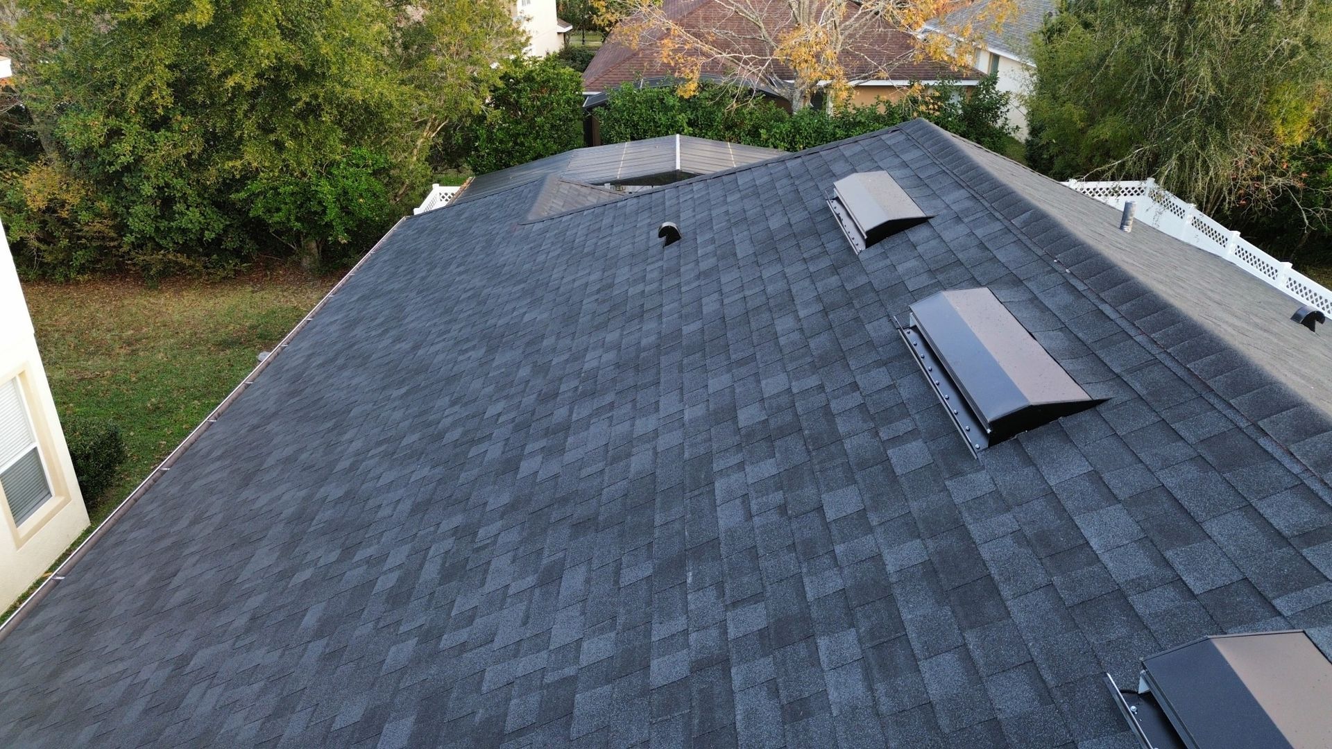 Roof covered in dark gray shingles with skylights, viewed from above in a wooded suburban area