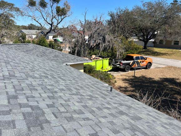 Roof shingles in foreground, with an orange utility vehicle and green dumpster in a yard under trees.