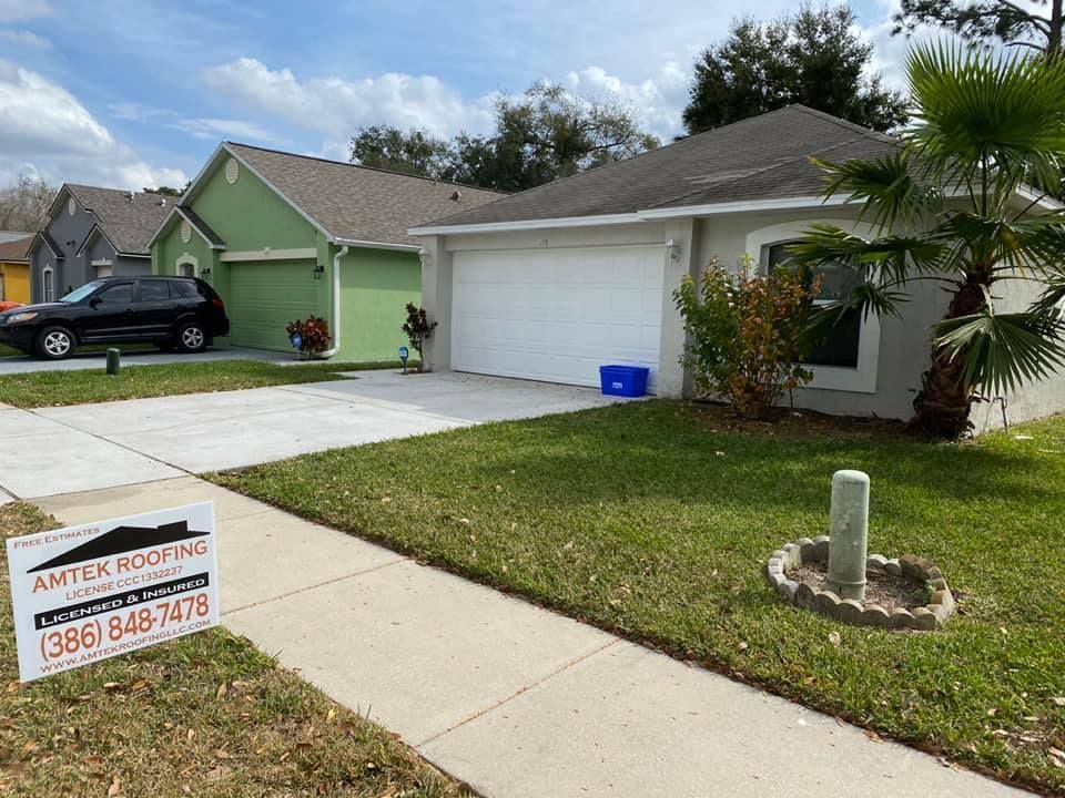 Suburban house with a white garage, green siding, driveway, lawn, and roofing company yard sign.