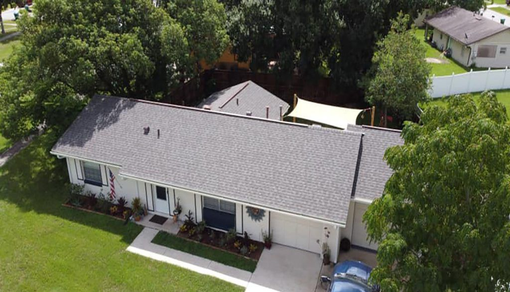 Aerial view of a single-story white house with a gray roof, surrounded by trees and a front lawn.