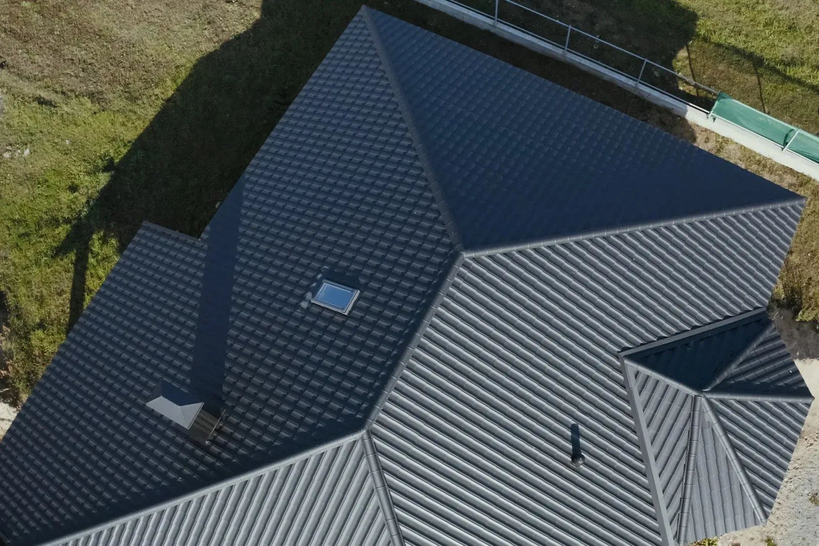 Aerial view of a dark gray shingled roof with multiple gables and skylights beside a grassy yard.