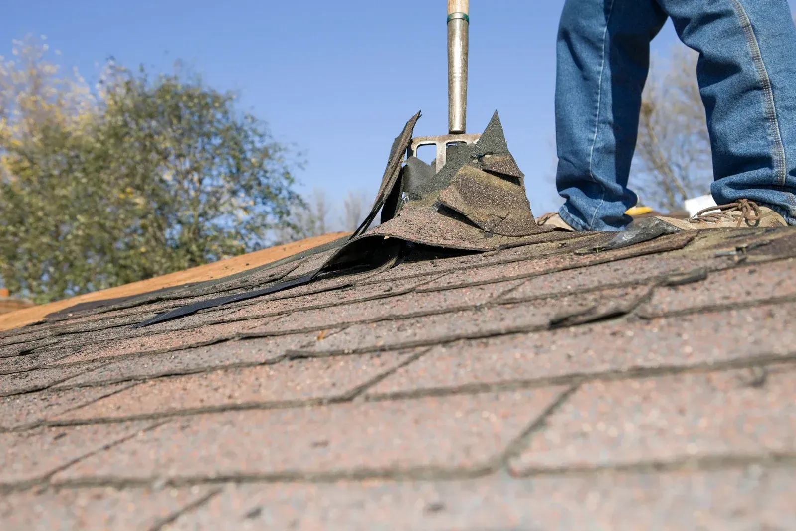 Worker in an orange shirt kneeling on a shingled roof using a tool under a blue sky