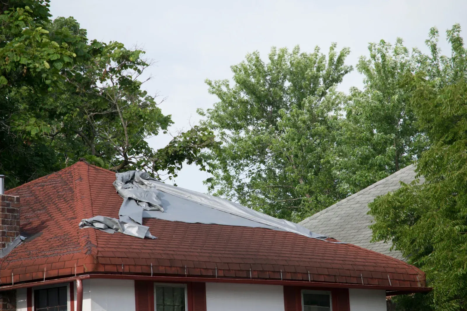 Storm-damaged red brick roof with torn shingles and tarp, surrounded by leafy trees
