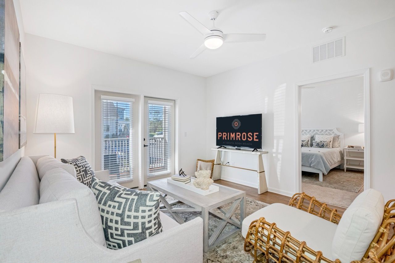 Bright living room in a white-themed apartment with a sofa, coffee table, and TV; doorway to a bedroom at Primrose at Santa Rosa Beach in Santa Rosa Beach, FL.