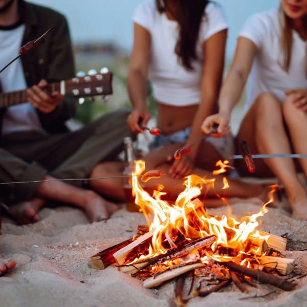 Three friends sit around a beach bonfire at twilight, roasting sausages on sticks while one plays an acoustic guitar.